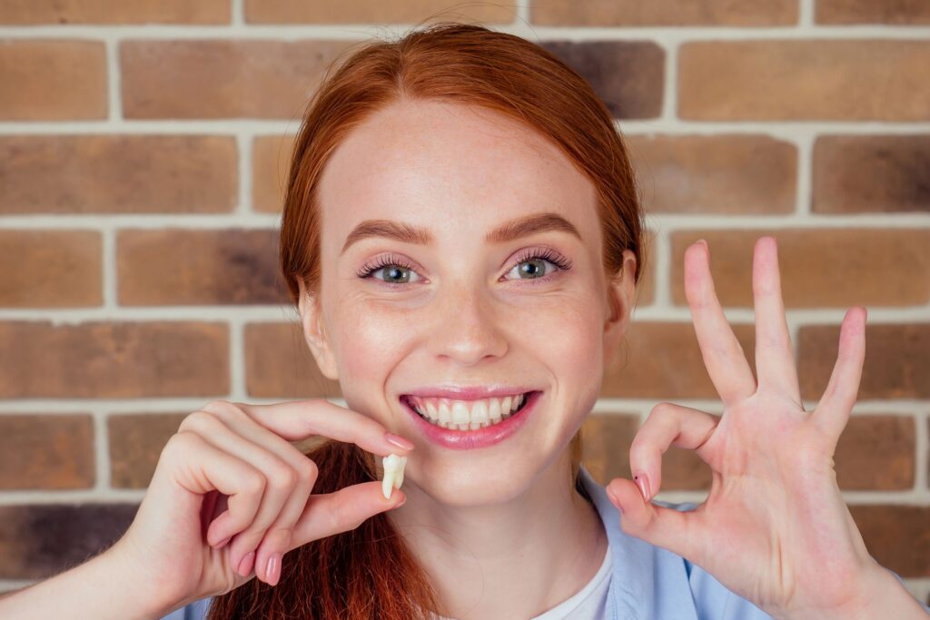 Woman with red hair holding extracted tooth and making ok sign with other hand